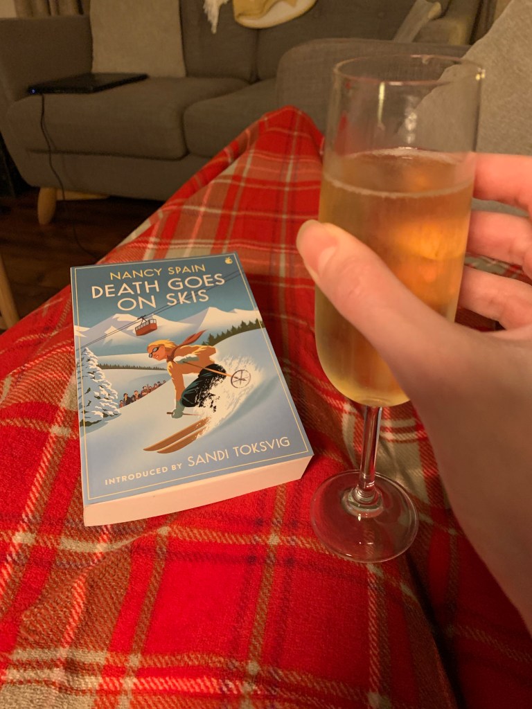 A copy of Death of Skis rests on a tartan rug while a hand holds a glass of champagne